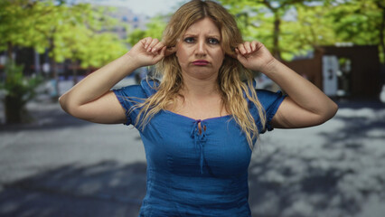 Woman wearing blue dress plugs ears with fingers on city street by trees and a building, sunlit...