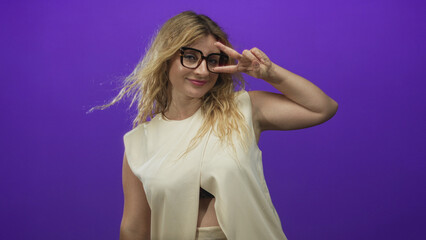 Woman with blonde hair wearing glasses smiling and making a peace sign in a purple studio with...