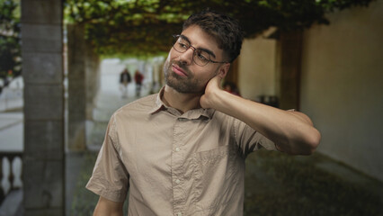 Man wearing glasses touches neck on street under leafy canopy and next to stone building facade;...