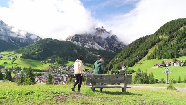 Peaceful walk in nature with view of Santa Maddalena, Val di Funes Elderly couple walking toward bench with scenic alpine village and snowy Dolomites in background