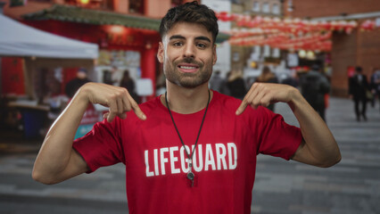 Fototapeta premium Hispanic man points finger to red lifeguard shirt and shows whistle on crowded city street; confidence.