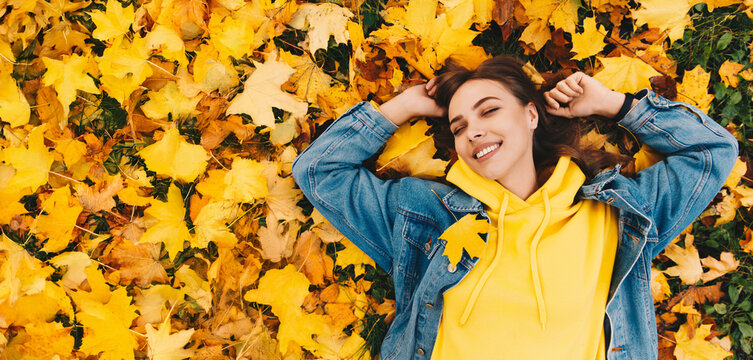Joyful young woman in yellow hoodie and denim jacket lying on golden autumn maple, leaves, smiling with closed eyes. Bright fall colors, cozy mood, and relaxed seasonal atmosphere. - Powered by Adobe