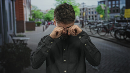 Man rubs eyes on narrow european street set against brick buildings and rows of parked bicycles;...