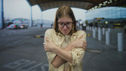 Fototapeta premium Young blonde woman shivering and hugging her arms at airport terminal outdoors; cold discomfort.