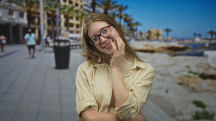 Fototapeta premium Young blonde woman wearing glasses laughs and covers mouth with hand on sunlit street by building; joy.