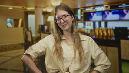 Woman with blonde hair wearing glasses stands with hands on hips beside tv display in restaurant;...