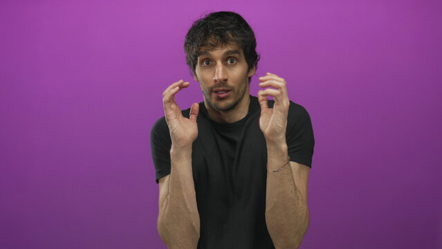 Young man raising hands defensively in studio with purple wall and tense expression and hunched posture; anxiety.
