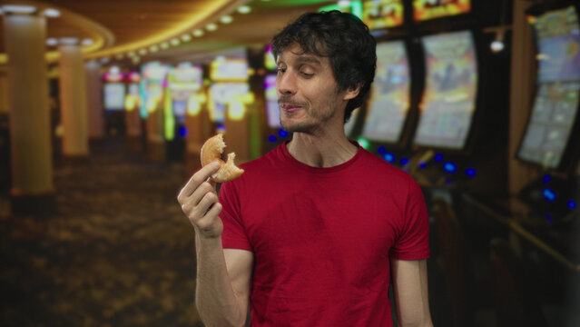 Man in red shirt holding donut chewing in casino against bright row of slot machine screens; satisfaction.
