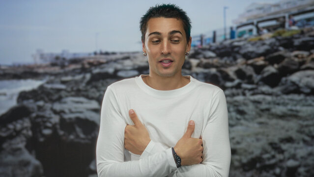 Young hispanic man standing outdoors by rocky seaside beach in casual white attire showing emotion. - Powered by Adobe