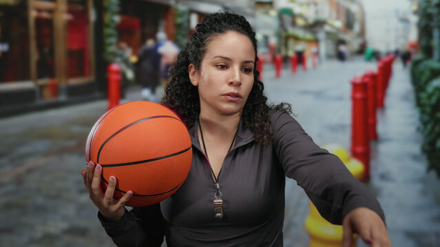 Woman holding a basketball on a bustling city street, showcasing an athletic and determined expression, with a focus on her confident stance and urban outdoor setting. - Powered by Adobe