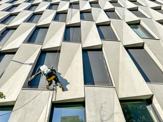 industrial climber, window cleaner works on office building at height of several floors on cables, washing windows of metropolis business center with mops and pressure water machines