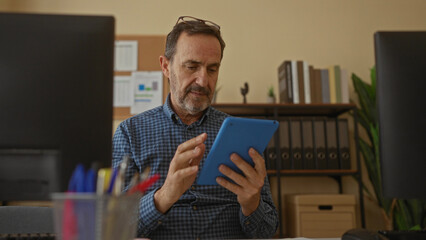 Man in office using tablet surrounded by computers and office supplies, wearing glasses and focused...