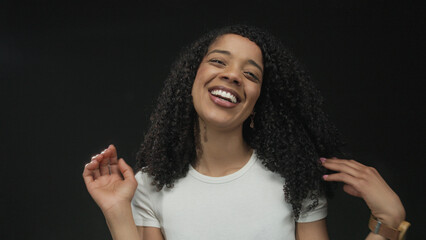 African american woman wearing white t shirt smiles and touches curly hair with both hands in...
