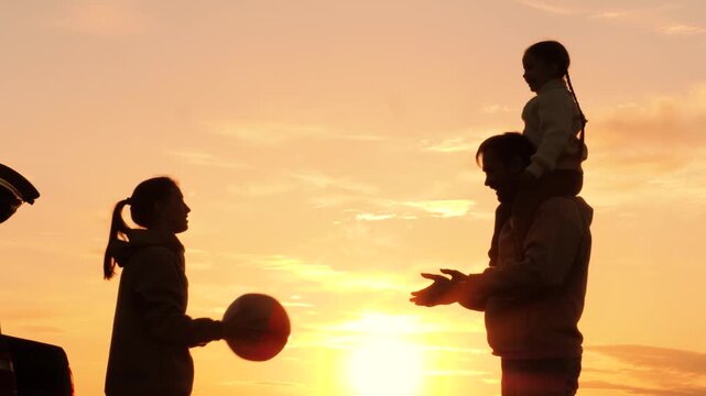 Family playing with ball in nature at sunset, silhouettes. Daughter sits on father's shoulders, throwing ball with mother. Family travel adventures by car, active leisure. Weekend road trip to nature.
