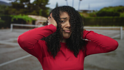 Woman with curly hair and red long sleeve shirt covers ears on outdoor tennis court painted court...