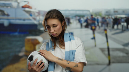 Woman holding soccer ball near port with boat in background, wearing scarf, outdoor setting...