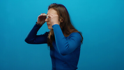 Woman peering through fingers on isolated blue background suggests curiosity, with a caucasian...