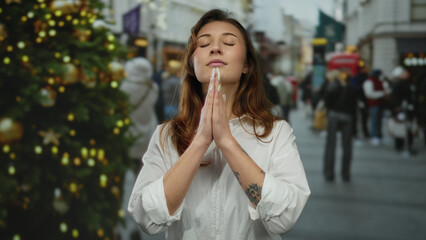 Fototapeta premium Woman praying outdoors on city street near christmas tree surrounded by people in festive setting wearing white shirt in urban area during holiday season.