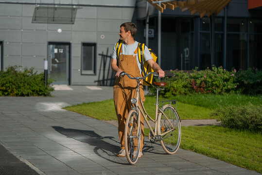 Man in work uniform walking his bicycle with a large delivery backpack on a sunny city sidewalk, urban bike courier delivering food and packages, side view, active and eco-friendly lifestyle