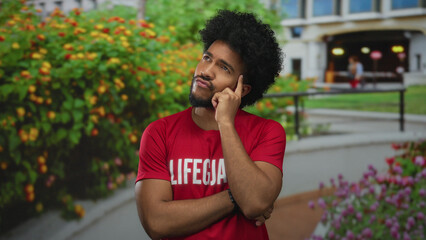 Man pondering outdoors with a thoughtful expression in a park setting wearing a lifeguard shirt,...