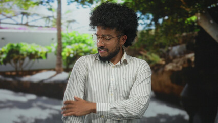Fototapeta premium Man wearing glasses and striped shirt stands outdoors with trees in the background, expressing thoughtful mood on a sunny day.