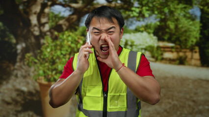 Naklejka premium Asian man shouting on smartphone outdoors wearing safety vest in park setting with trees and plants visible in background.