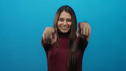 Woman smiling and pointing forward against a vibrant blue background wearing a maroon sweater...
