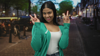 Woman wearing green cardigan showing hand peace sign with raised arms on busy city street at dusk;...