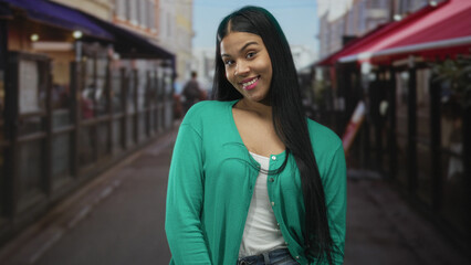 Latin woman with long dark hair in green cardigan smiles on city street outside cafe; confidence...