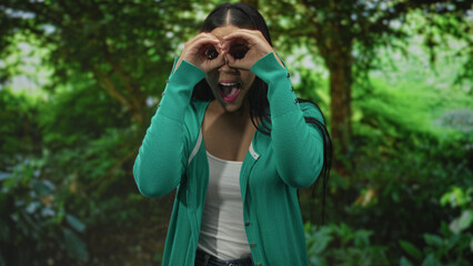 Woman makes binocular gesture with hands over eyes amid dense green forest trees; adventure...