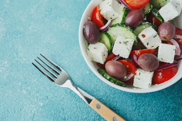Greek salad with feta cheese in white bowl and fork on blue background