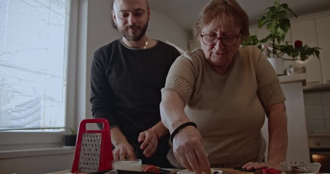 A senior lady and a man prepare a meal together in the kitchen. They add cheese and other toppings to a baking sheet.