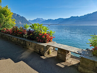 Sunny day on the Montreux lakeside promenade, Switzerland, with colorful flowers, a stone bench, and a calm view of Lake Geneva under a clear blue sky.