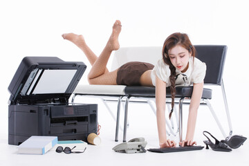 Asian pretty young woman confident braided hair in office costume lying on bench, poses playfully beside large printer books sunglasses keyboard landline phone shoes in white studio fashion setting.