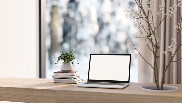 White screen computer and books with dried branch through wooden table and snow outside glass wall.