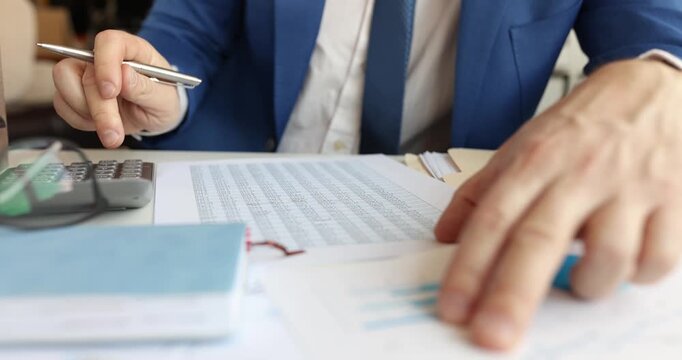 Man in suit studies printed accounting report on desk with pen. Analyst marks figures highlighting rows and checking totals for update sheet