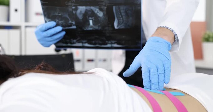 Radiologist positions x-ray image over patient lower back with kinesio tape. Specialist studies spine picture evaluating lumbar area carefully