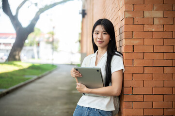 Young asian woman student holding a laptop while standing leaning against brick wall of a building.