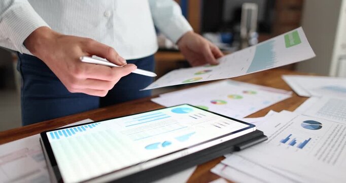 Businesswoman compares data on digital tablet and paper in office. Female entrepreneur analyzes business activities and trends with diagrams