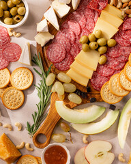festive charcuterie board with assorted sliced salami, cheese, olives, crackers, grapes, nuts, honeydew melon, apple wedges, rosemary, and dipping sauce on light stone table,