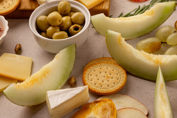 close-up festive snack platter with green olives, cheese wedges, brie, honeydew melon slices, fresh grapes, round crackers, apple slices and pistachios on rustic stone table, gourmet