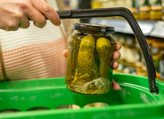 Woman buys pickles in jar while putting them in shopping cart at grocery store.