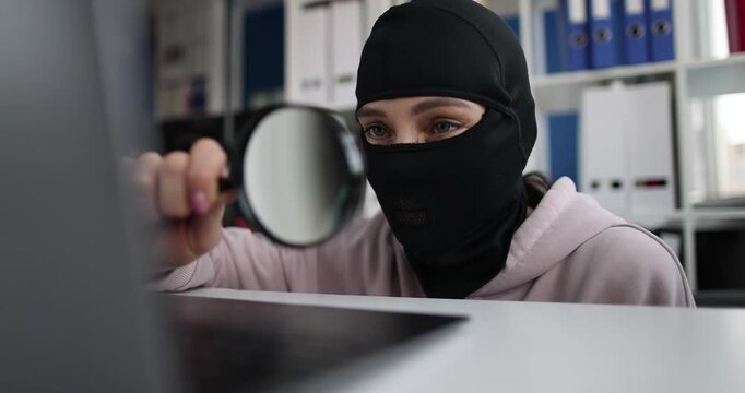 Female thief in ski mask looks at laptop screen through magnifying glass in office. Criminal steals confident data from computer in corporate setting