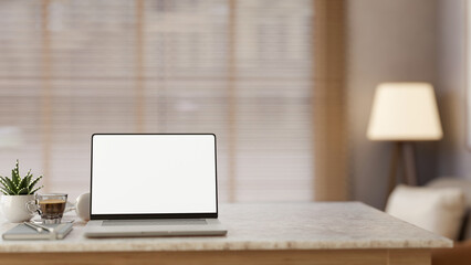 Fototapeta premium Blank screen laptop with book and coffee on marble table in a blurred living room with lamp and sofa