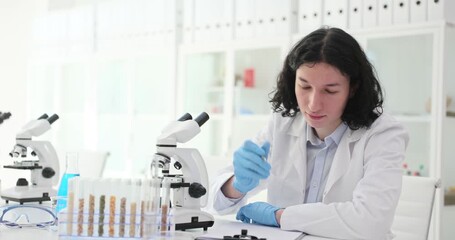 Young man lab assistant examines cereals with microscope in laboratory. Scientist marks notes of agricultural plants diseases in lab setting - Powered by Adobe