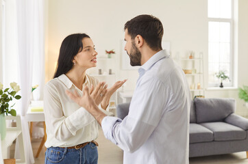 Fototapeta premium Young couple man and woman standing at home and arguing shouting in quarrel. Angry unhappy husband and wife quarreling in the living room. Marriage problems, relationship and divorce concept.