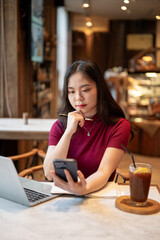 Asian woman holding pen under chin looking at smartphone aside laptop and coffee at table in a cafe.