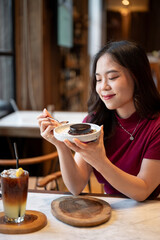 Young asian woman holding spoon and chocolate cake on plate aside orange coffee at table in the cafe