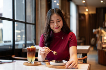 Young asian woman holding spoon eating chocolate cake aside orange coffee cup at table in the cafe.