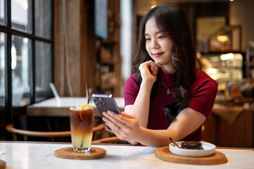 Young asian woman looking at phone or taking picture of orange coffee aside cake on table in a cafe.